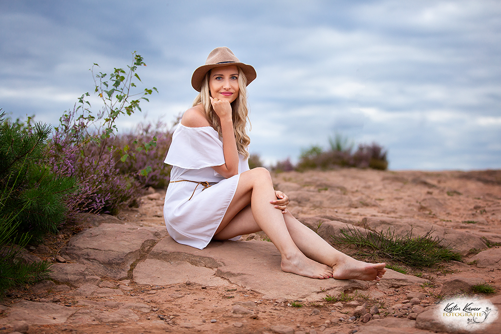 Frau im weißen Kleid mit Hut sitzt auf einem Felsen in einer natürlichen Umgebung.