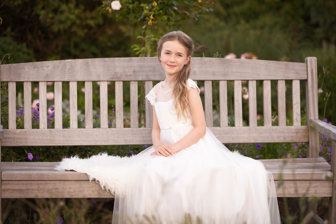 Mädchen in weißem Kleid sitzt auf einer Holzbank umgeben von Blumen.