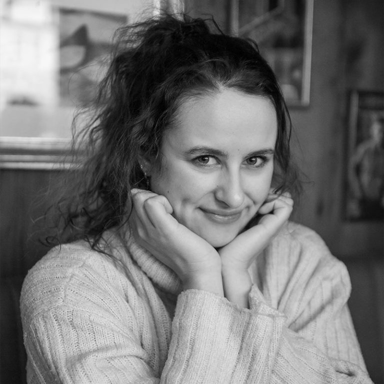 Momentaufnahme im Cafe Eine lächelnde Frau mit lockigem Haar, die freundlich die Hände unter dem Kinn stützt.